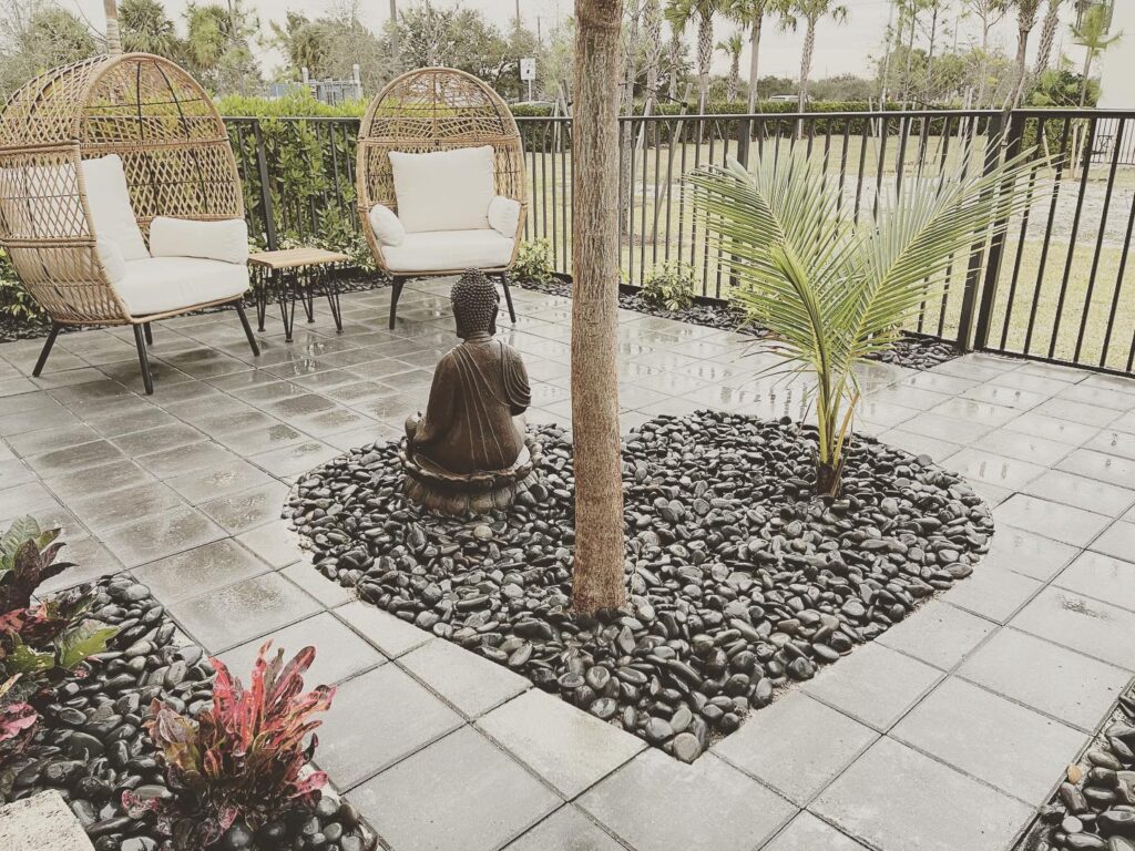 A tranquil outdoor space featuring two wicker chairs, a small table, and a Buddha statue surrounded by black pebbles and greenery, evoking calmness.