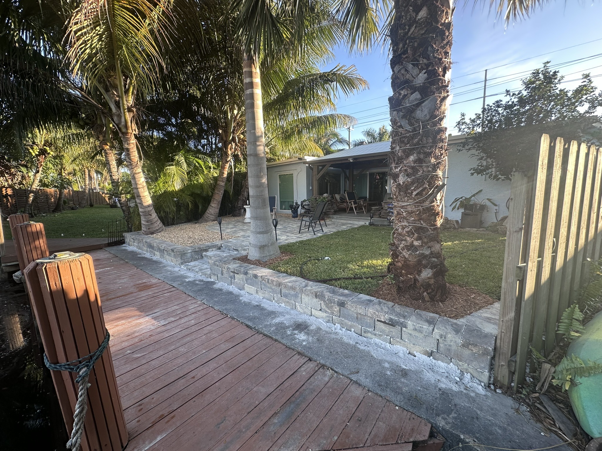 Tropical backyard scene featuring palm trees, a stone patio, and outdoor seating. A wooden dock leads to a tranquil waterway, enhancing the relaxing atmosphere.