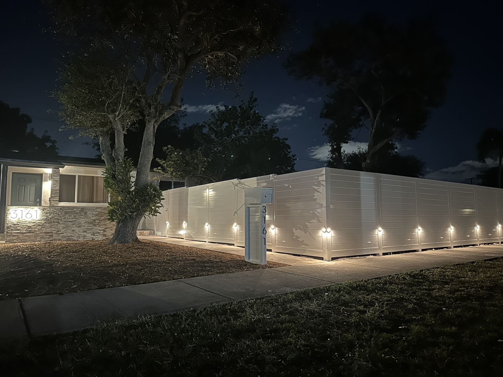 Modern home exterior lit at night, featuring a stone facade, lush greenery, and a secured white fence. The address "3161" is prominently displayed.
