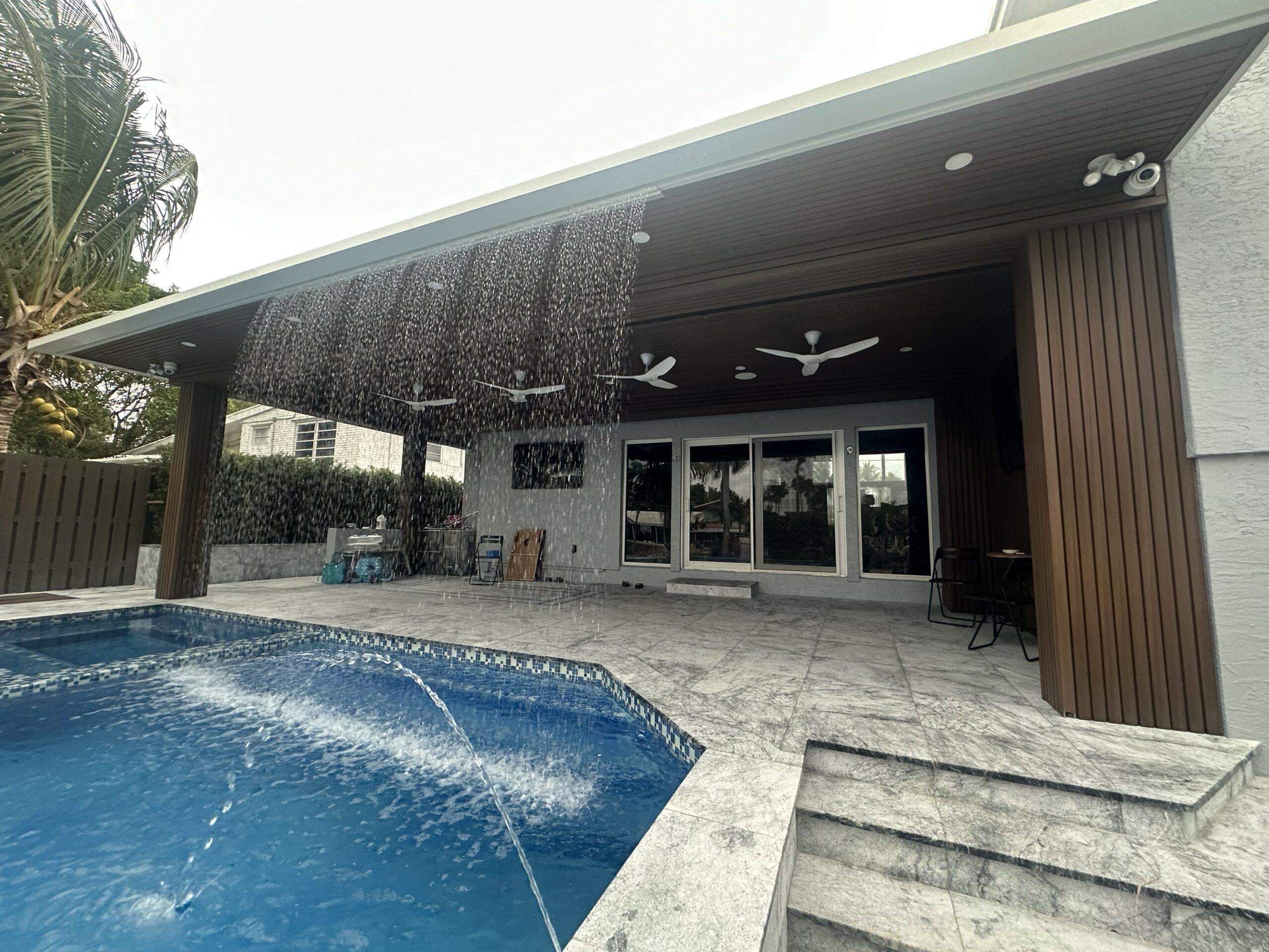 Modern outdoor patio featuring a pool with water fountains, under a shaded area with ceiling fans and spacious seating, ideal for relaxation.