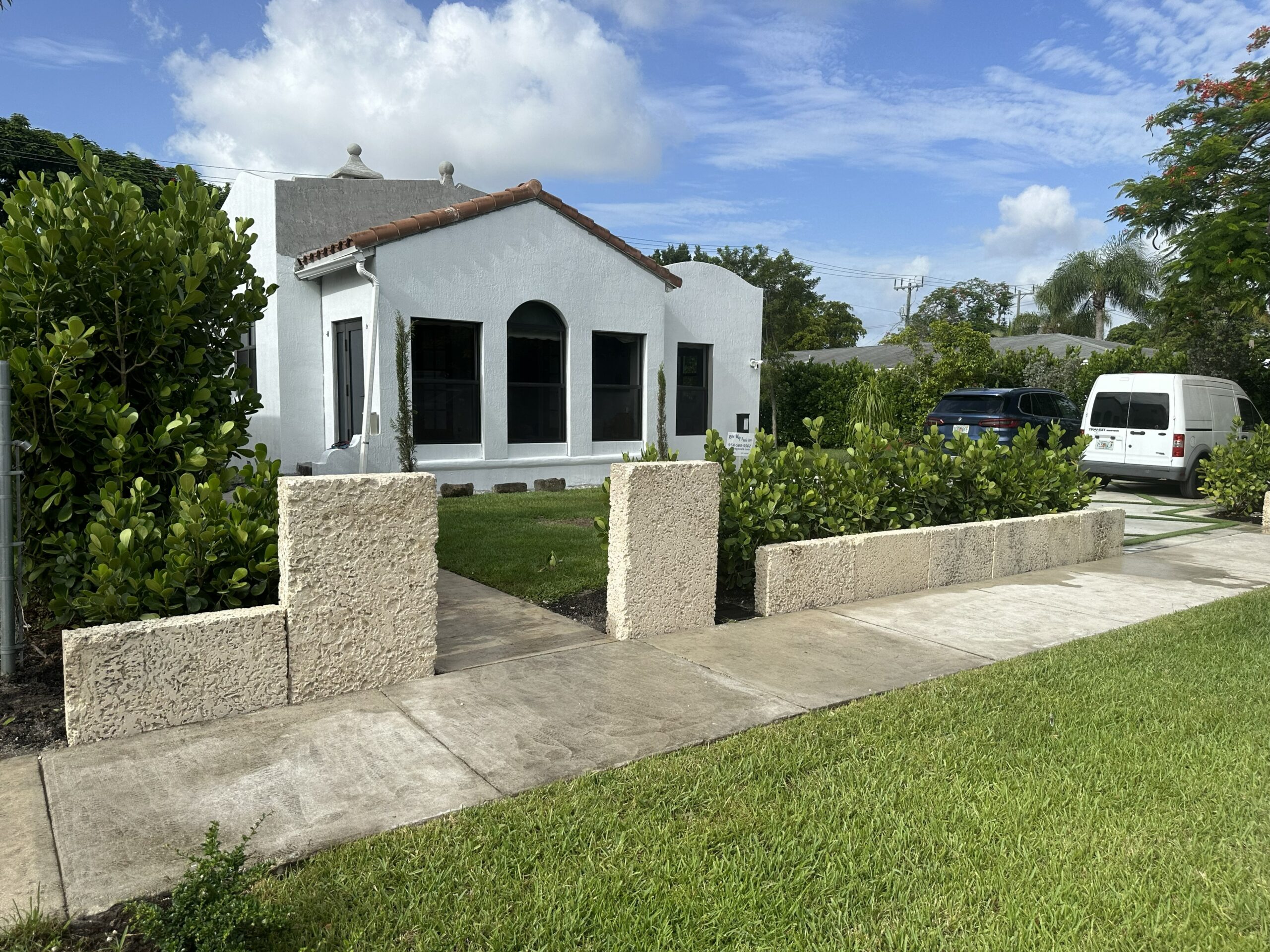 White stucco house with arched windows and clay tile roof, surrounded by greenery, with a concrete sidewalk and parked vehicles.