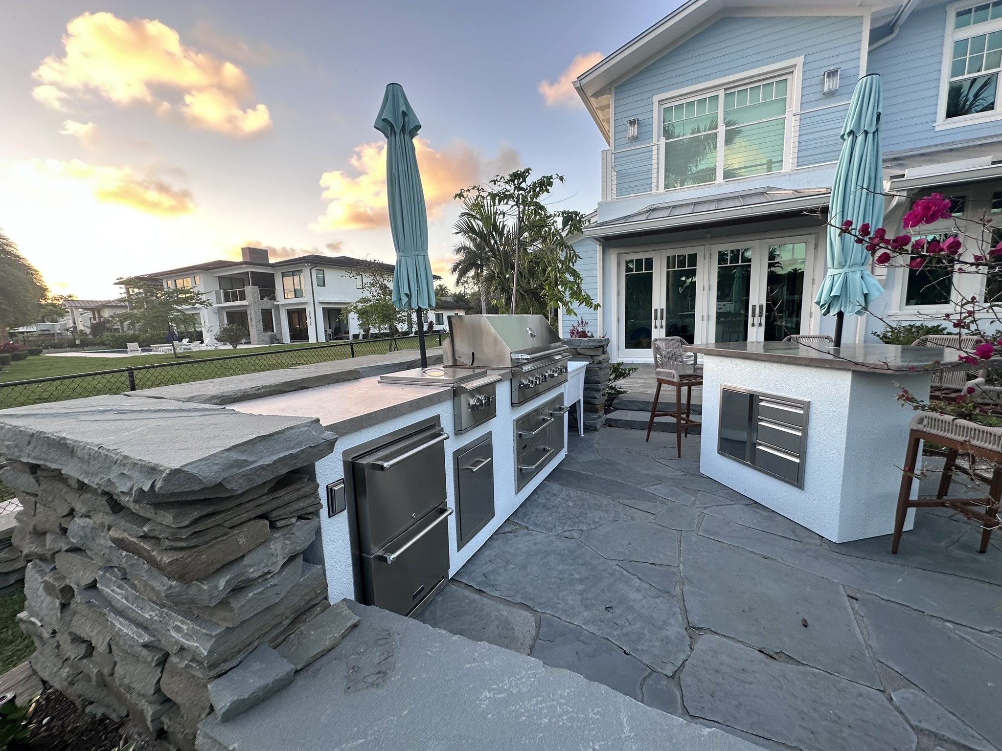 Outdoor kitchen featuring a stainless steel grill, stone countertops, and seating, set against a modern home with a sunset backdrop.