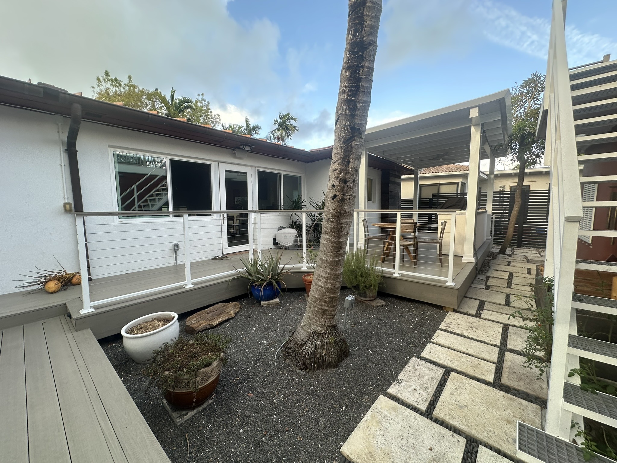 A modern patio area featuring a wooden deck, railing, and landscaped paths, with palm trees against a cloudy sky, creating a serene outdoor space.