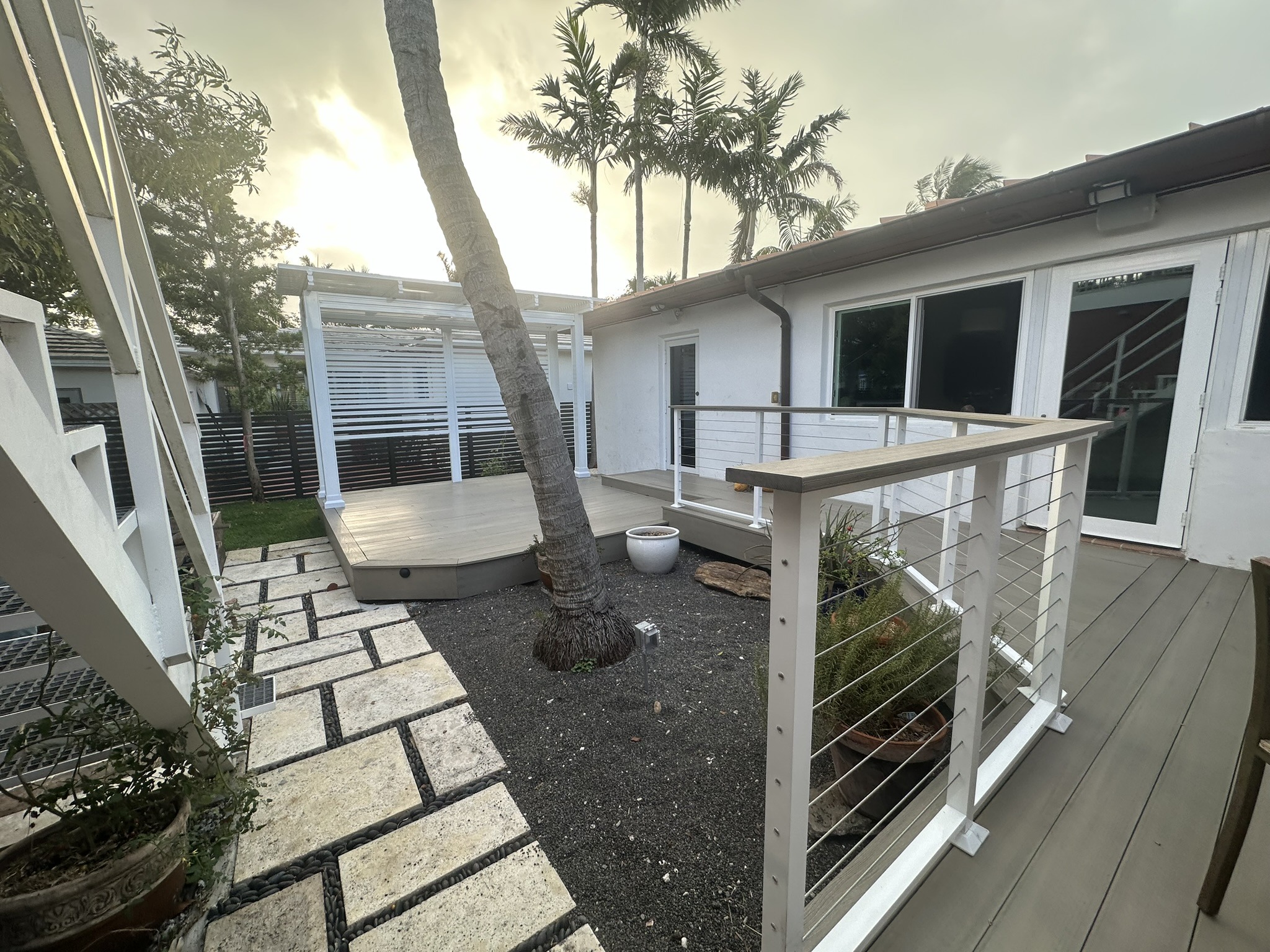 A modern patio area featuring a wooden deck, railing, and landscaped paths, with palm trees against a cloudy sky, creating a serene outdoor space.